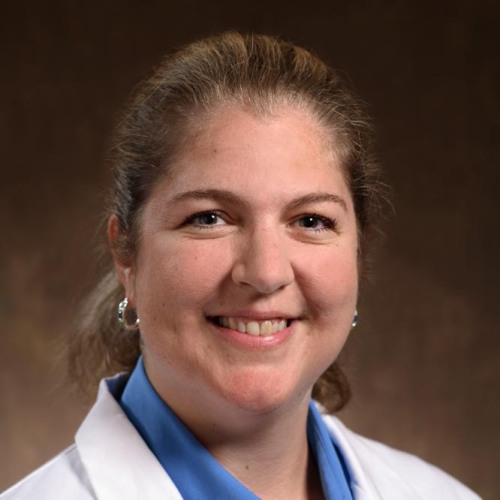 woman smiling wearing a lab coat and a blue shirt against a brown background