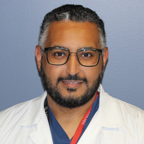 man wearing blue scrubs, glasses, a lab coat and a red lanyard