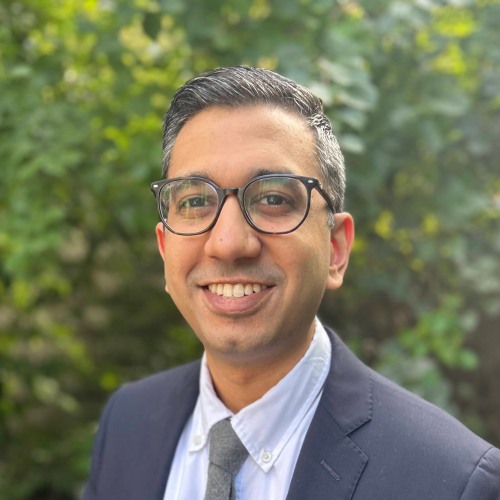 man wearing glasses and a suit smiling against a green leafy background