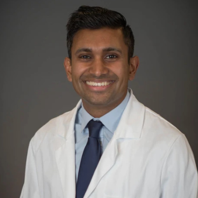 man wearing a lab coat, blue shirt and blue time smiling against a grey background