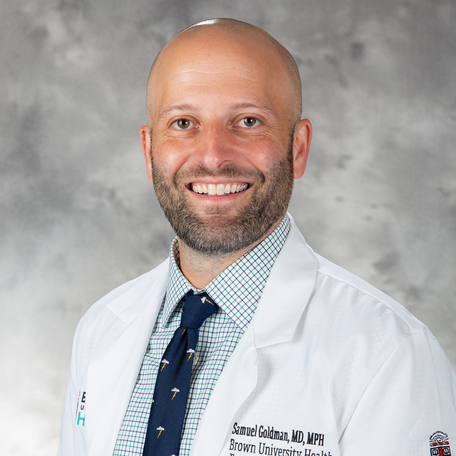 man wearing a lab coat, blue shirt and blue tie smiling against a grey background
