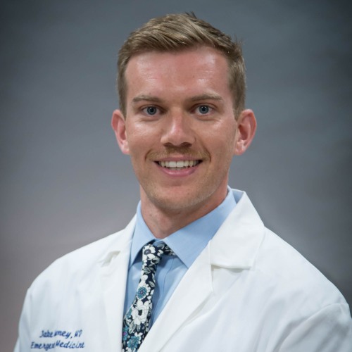man wearing a lab coat, blue shirt and blue tie smiling against a grey background