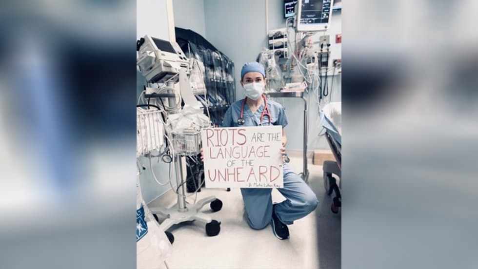 person kneeling and holding a sign that says "Riots are the language of the unheard"